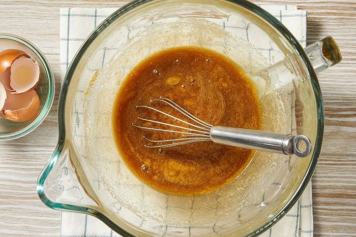 Overhead horizontal image of wet ingredients being mixed in a clear glass bowl with a metal whisk. A smaller bowl of empty egg shells sits beside the mixing bowl, on a wooden surface with a patterned towel.