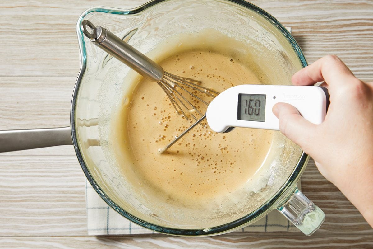Overhead horizontal image of batter in a clear glass mixing bowl. A hand is using a Thermapen to check the temperature of the batter, while a whisk rests inside.