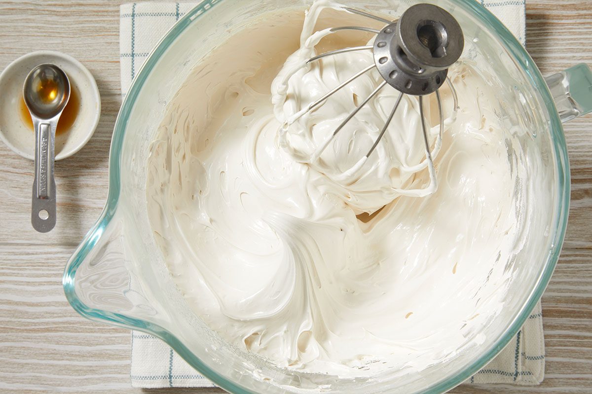 Overhead horizontal image of a large glass mixing bowl containing white whipped cream with a metal whisk inside. A small white dish with a spoonful of vanilla extract sits to the side, on a light wood surface.