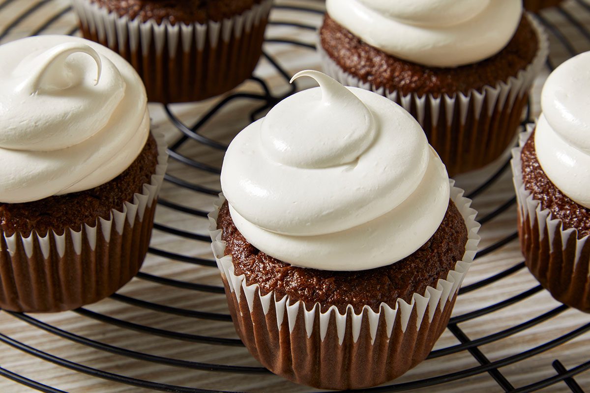 Eye Level horizontal image of chocolate cupcakes with white frosting on a black cooling rack. The cupcakes are in white paper liners and have a swirl of white frosting on top.