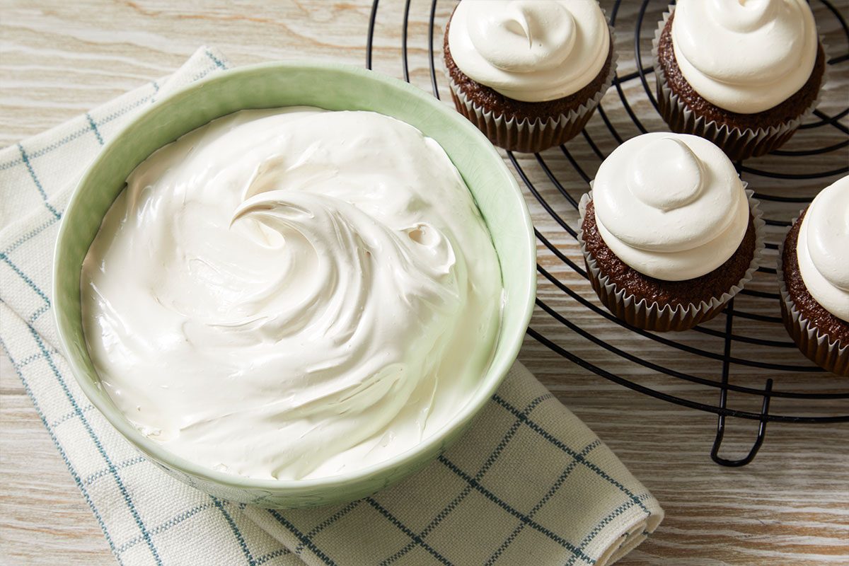 Overhead horizontal image of a light green bowl filled with smooth white frosting. To the right are cupcakes topped with the same frosting, on a black wire rack.