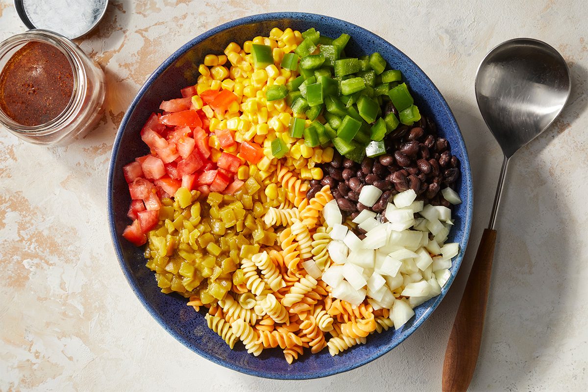 Overhead horizontal image of Southwest Corn Salad ingredients arranged in sections within a blue bowl. Visible are 3 cups cooked tricolor spiral pasta, 1 can (15-1/4 ounces) whole kernel corn, drained, 1 can (15 ounces) black beans, rinsed and drained, 1 large tomato, seeded and chopped, 1/2 cup chopped onion, 1/2 cup chopped green pepper, 1 can (4 ounces) chopped green chiles. A jar of dressing and a spoon are also visible. | [Original] RMS 22213 Southwest Pasta Salad; How to; Step 1 In a large bowl; combine the first 7 ingredients. In a jar with a tight-fitting lid; combine oil; vinegar; sugar; chili powder; salt and cumin; shake well.