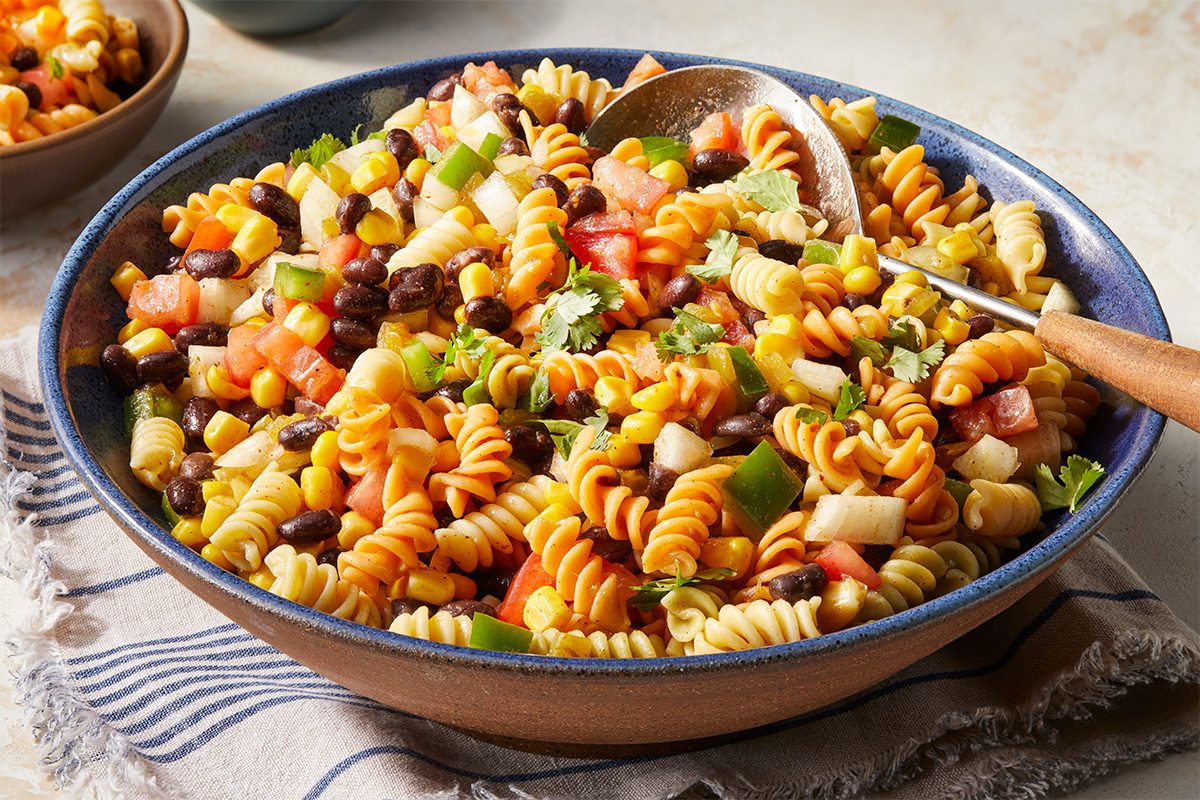 Eye Level horizontal image of Southwest Corn Salad in a blue ceramic bowl with a serving spoon. The salad contains tricolor spiral pasta, corn, black beans, tomato, onion, green pepper, and cilantro. | [Original] RMS 22213 Southwest Pasta Salad; Horizontal AP; Pull