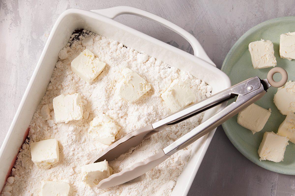 A pair of metal tongs picking up a cube of cheese or tofu from a baking dish filled with flour, with more cubes on a green plate nearby.
