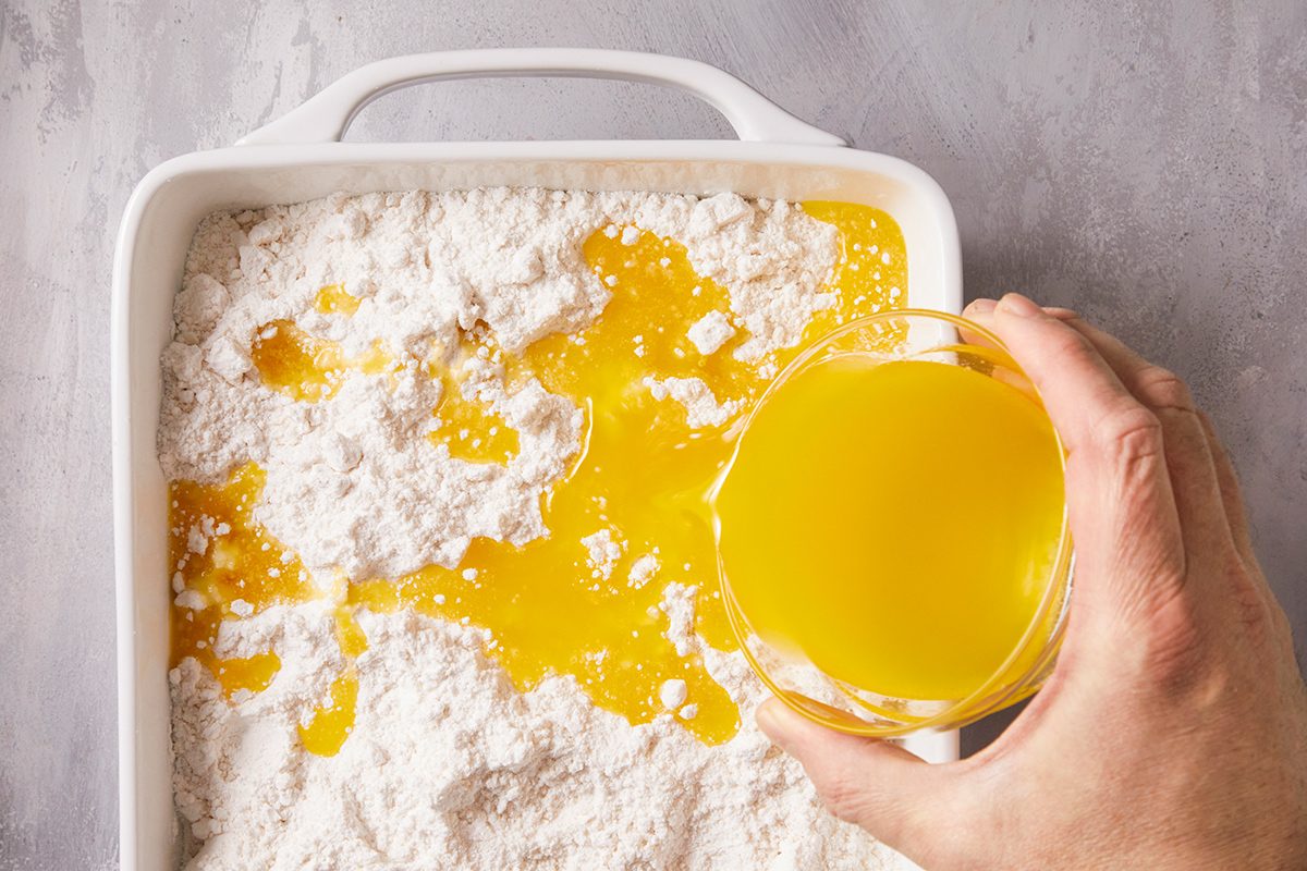 A hand pours melted butter over a layer of dry cake mix in a white baking dish, preparing ingredients for baking.