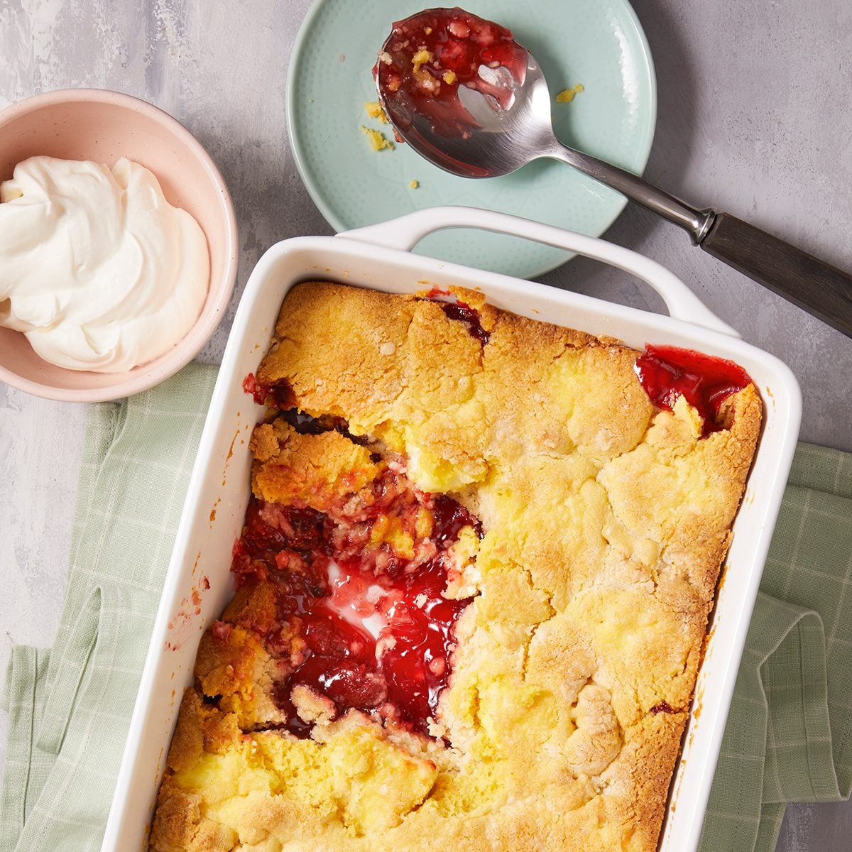 A rectangular baking dish of fruit cobbler with a golden, crumbly crust and a portion missing, revealing a red fruit filling. Nearby are a spoon with filling residue and a bowl of whipped cream.