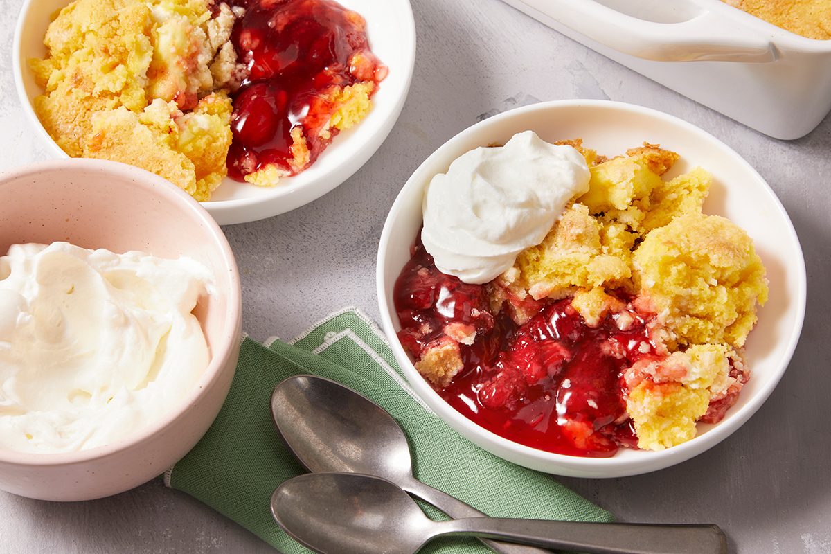 Two bowls of strawberry cobbler topped with whipped cream are on a table, with an extra bowl of whipped cream and two spoons on a green napkin nearby. A baking dish with more cobbler is in the background.