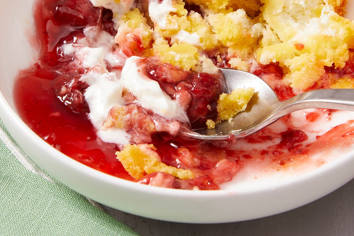 A close-up of a spoon scooping a dessert with red fruit filling, crumbly yellow cake, and whipped cream in a white bowl, with some dessert spread on a green napkin underneath.