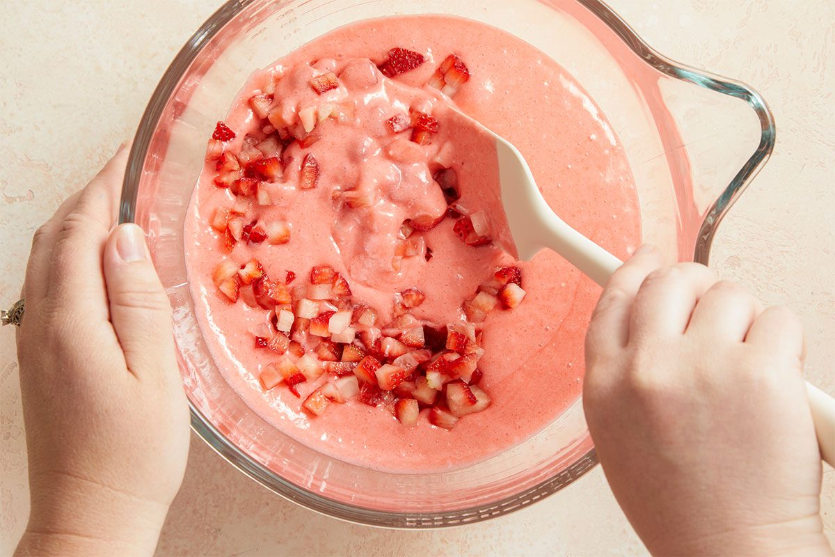 Overhead horizontal image of Strawberry Sheet Cake batter with chopped strawberries being mixed in with a white spatula in a clear glass bowl. The batter is pink in color and the ingredients are being combined by hand.