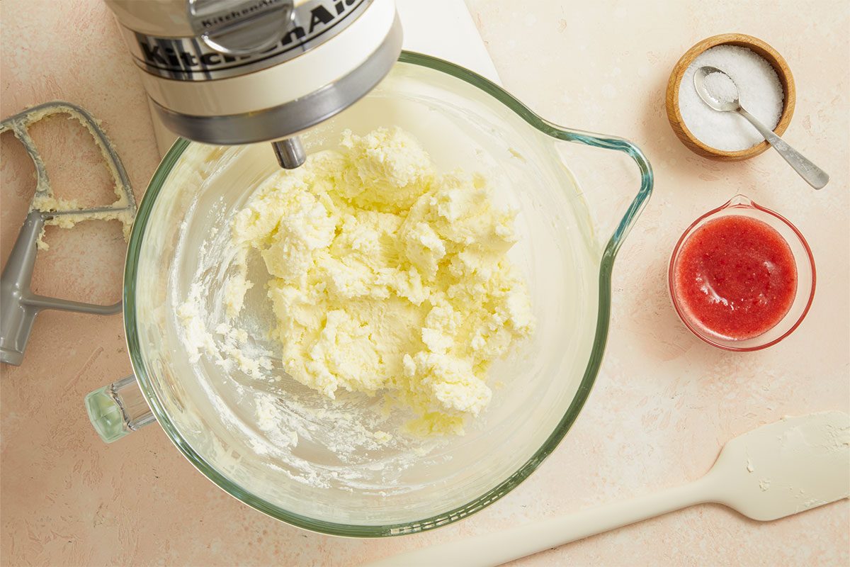 Overhead horizontal image of Strawberry Sheet Cake showing a KitchenAid mixer in a glass bowl of creamed butter and mascarpone cheese. Also visible are a bowl of fresh strawberries, sugar and a spatula.