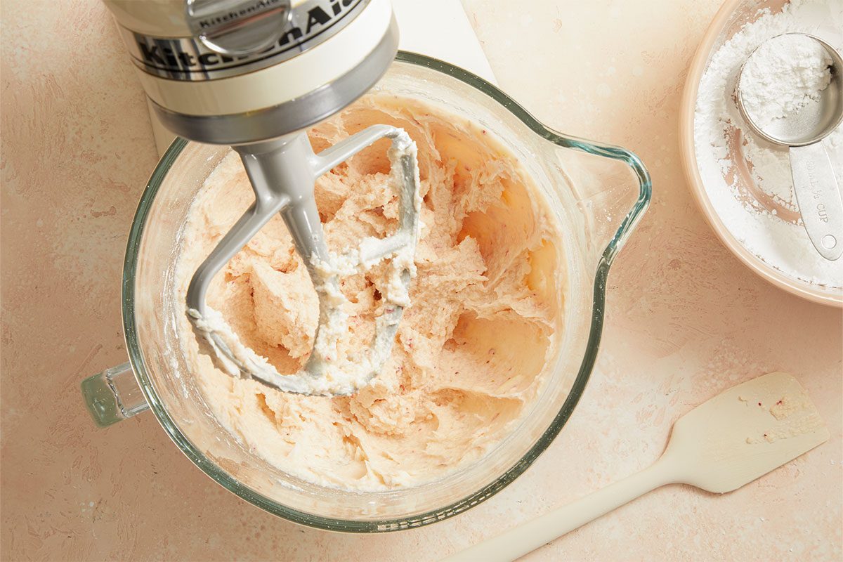 Overhead horizontal image of Strawberry Sheet Cake batter being mixed in a clear glass bowl by a KitchenAid mixer. Nearby, a bowl with confectioners' sugar and a measuring cup are visible.