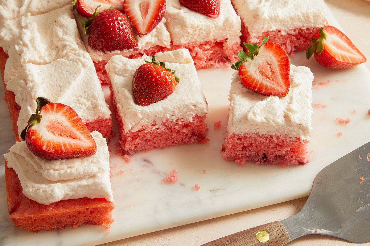 Eye Level horizontal image of Strawberry Sheet Cake cut into squares, topped with white frosting and sliced fresh strawberries, resting on a marble serving board. A spatula with a wooden handle is partially visible on the lower right.