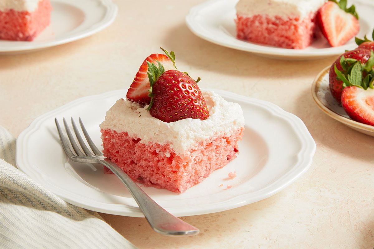 Eye level horizontal image of Strawberry Sheet Cake. One slice of cake is on a white plate with a fork next to it, topped with frosting and fresh strawberries, other slices of cake are in the background on other plates.