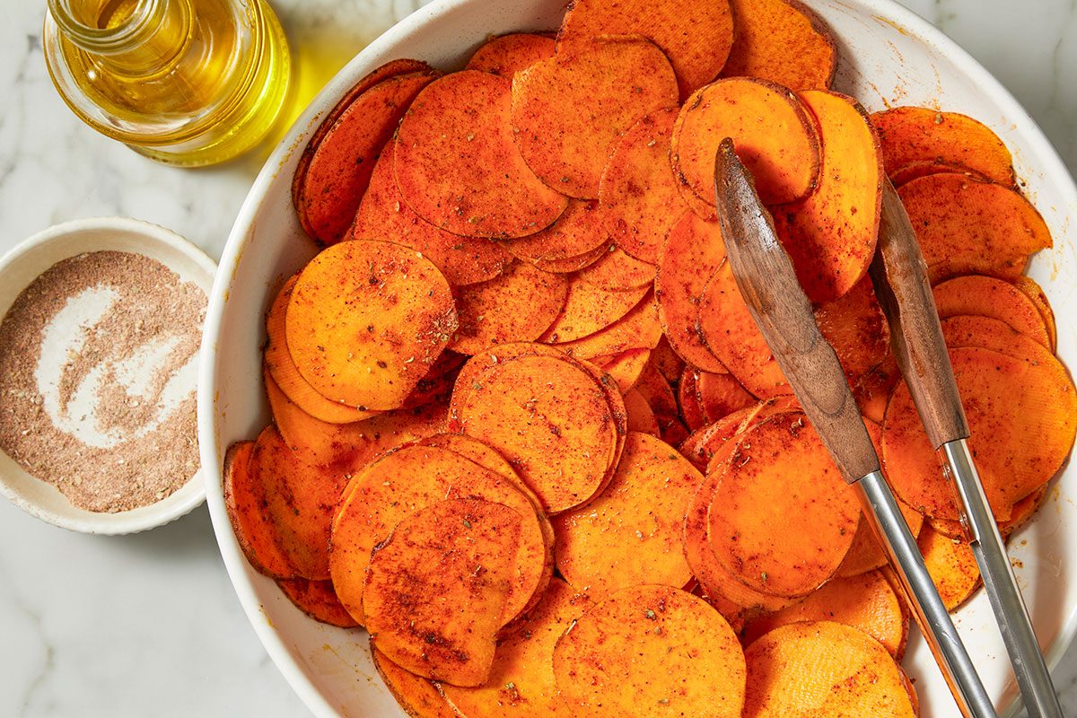 Overhead horizontal image of sliced sweet potatoes in a white dish, seasoned and ready to be cooked. Tongs lay across the potatoes, with a small bowl of seasoning and a bottle of oil in the background. | [Original] Overhead horizontal image of sliced sweet potatoes in a white baking dish. The potatoes are seasoned with spices and tossed with oil before roasting, with a bowl of seasoning and a bottle of oil nearby.