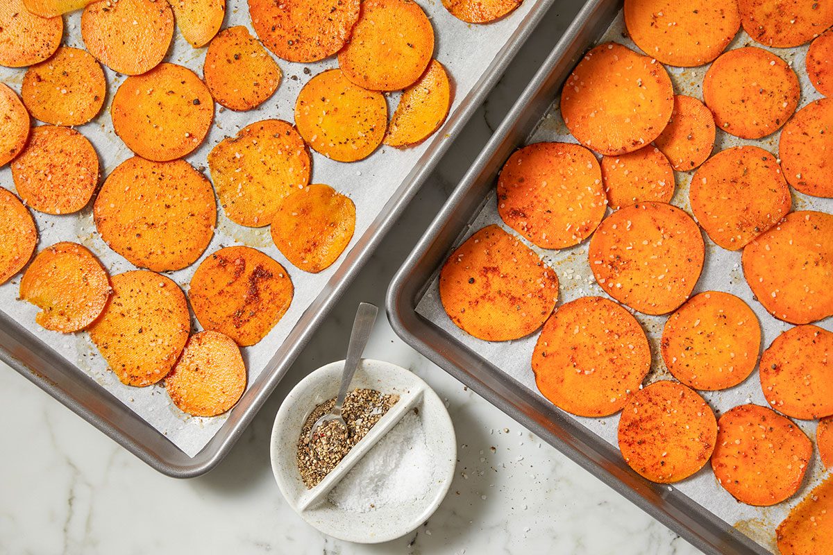 Overhead horizontal image of two trays of sliced sweet potatoes, seasoned and ready to be cooked. Salt and pepper are visible in a small bowl with a spoon, next to the trays.