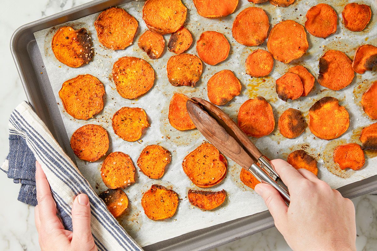 Overhead horizontal image of cooked sweet potato rounds on a baking sheet lined with parchment paper being lifted with tongs. A dishtowel is visible on the left.
