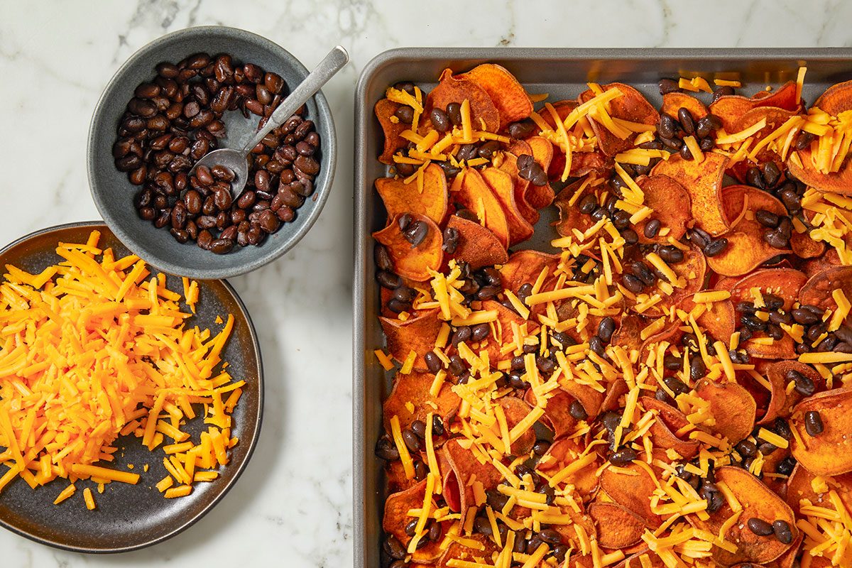 Overhead horizontal image of sweet potato chips being prepared on a baking sheet. Shredded cheese and black beans are being added to the chips before baking, with a bowl of beans and a pile of cheese on a plate to the left.