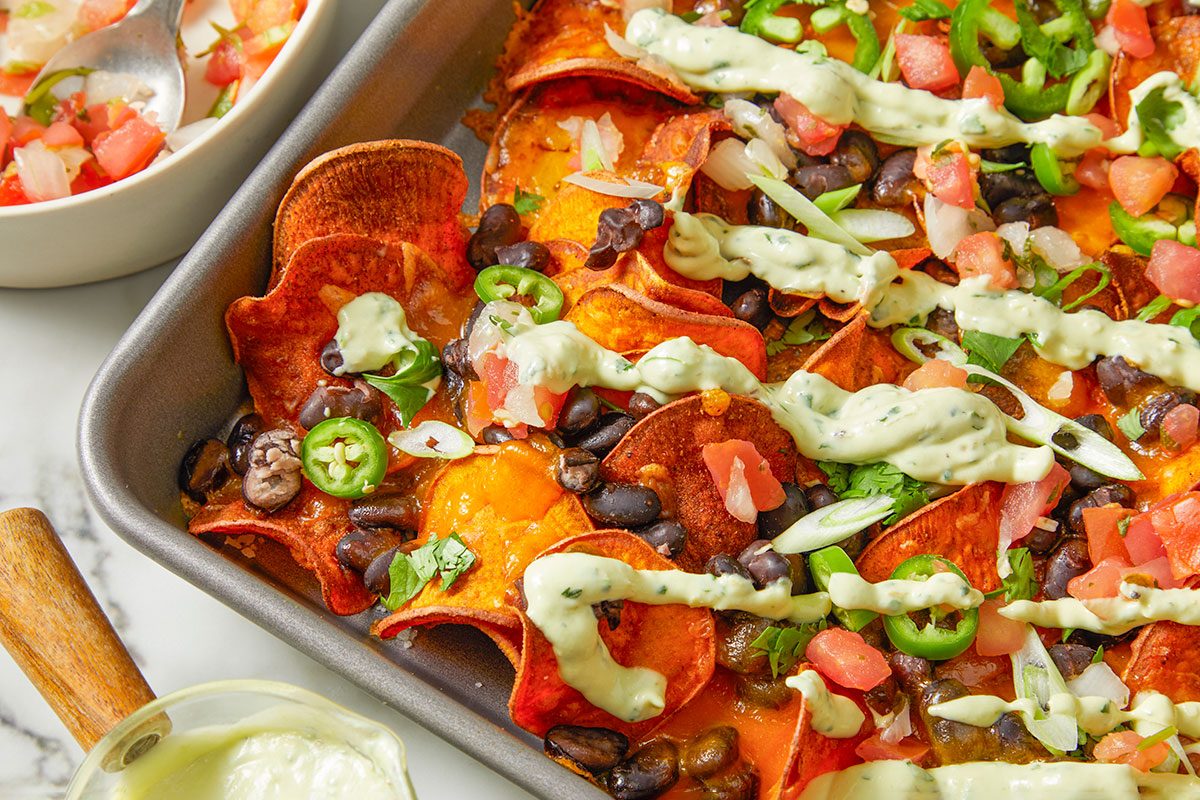 Overhead horizontal image of loaded sweet potato nachos. The sheet pan of chips is topped with black beans, tomatoes, green onions, and sliced jalapeños, and drizzled with a cream sauce. A bowl of tomato salsa and small pitcher of sauce are also in the shot. | [Original] Eye level horizontal image of loaded sweet potato nachos in a metal baking pan. The nachos are topped with black beans, jalapenos, chopped tomatoes and onions, cilantro, green onions, and a creamy sauce.