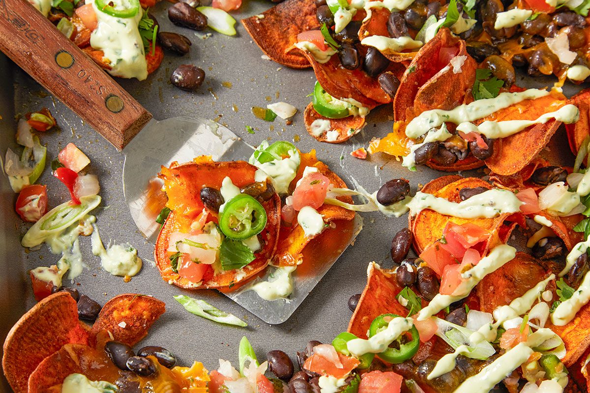 Overhead horizontal image of sweet potato nachos on a gray metal pan being lifted with a spatula. The nachos are topped with black beans, tomatoes, jalapenos, green onions, cilantro and a creamy white sauce.