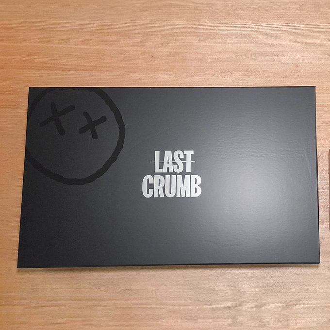 A black rectangular box with the words "LAST CRUMB" in bold white letters at the center and a doodle of two X’s inside a circle on the top left corner, placed on a wooden surface.