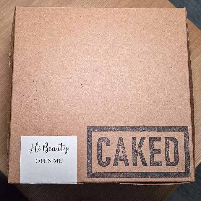 A brown cardboard box on a wooden surface with a label that says "Hi Beauty OPEN ME" and a large stamp reading "CAKED" on the lid.