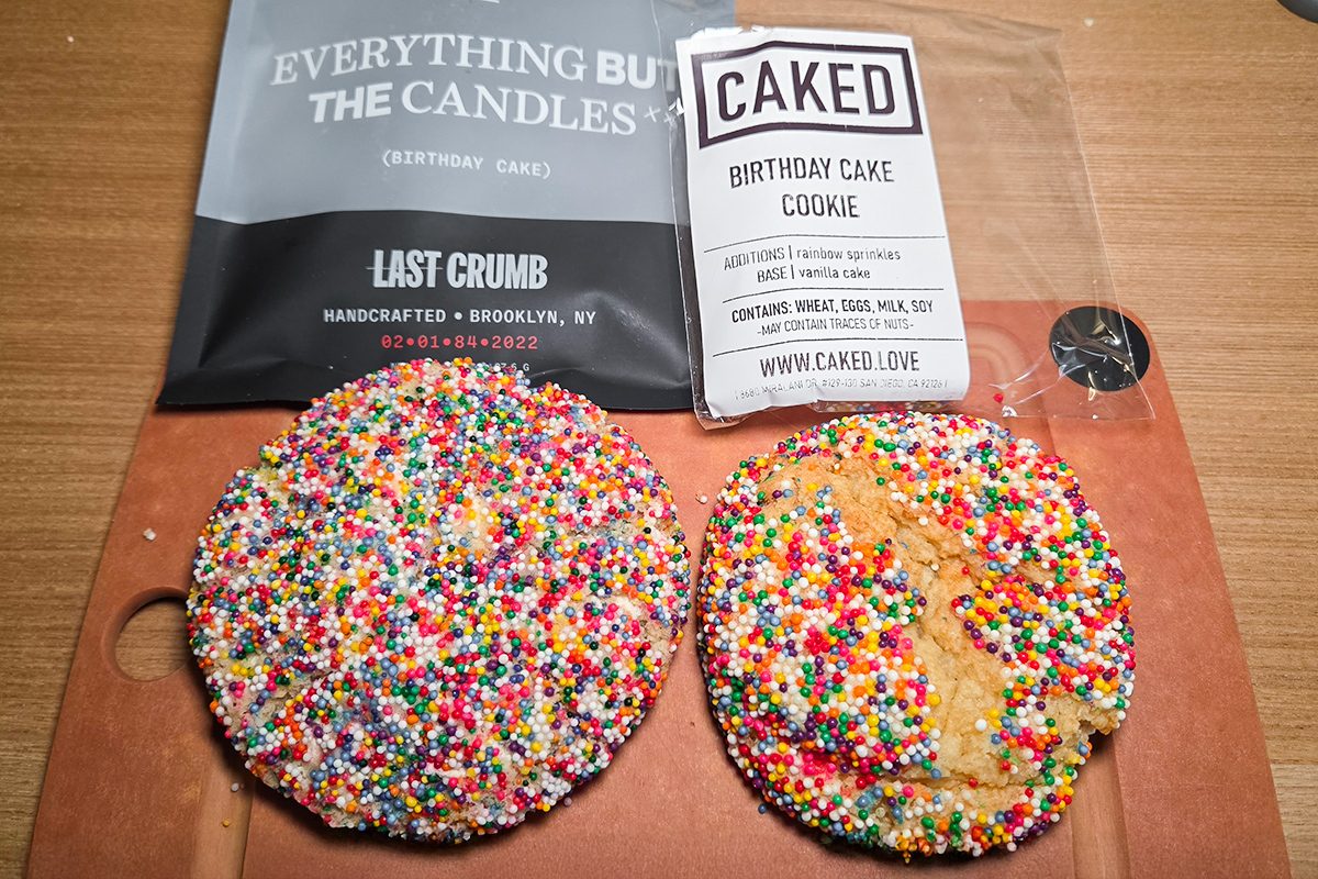 Two large cookies covered in colorful sprinkles sit on a wooden surface. Behind them are packages labeled "Everything But The Candles" from Last Crumb and "Birthday Cake Cookie" from Caked.
