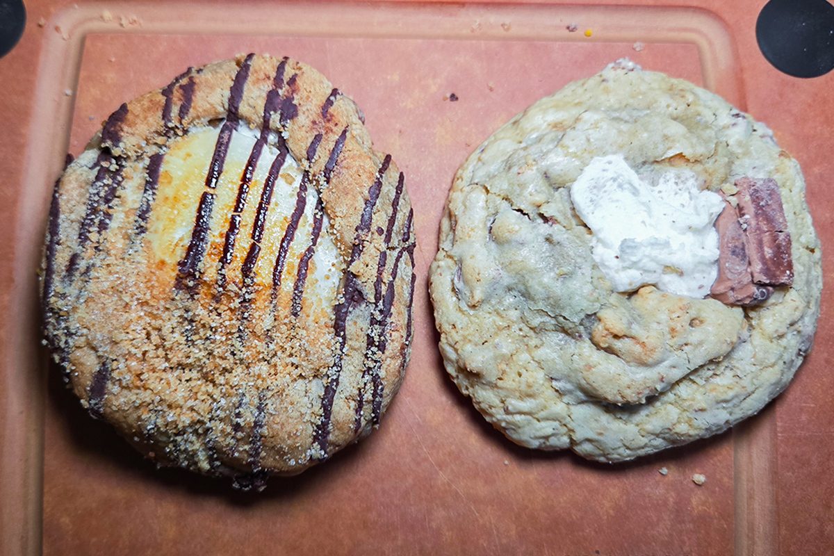 Two large cookies on a brown cutting board. The left cookie is topped with chocolate drizzle and crumbs, while the right cookie appears chunky with bits of marshmallow and chocolate.
