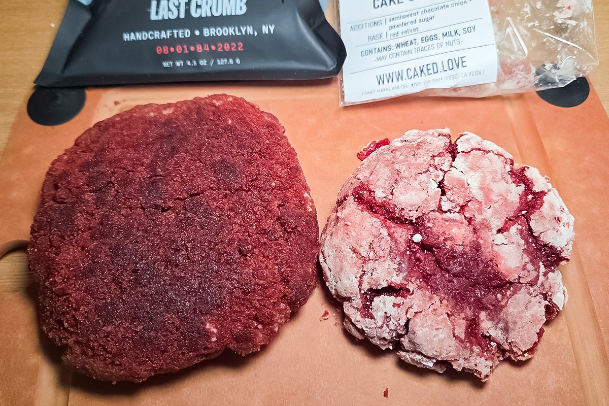 Two red velvet cookies on a brown surface; one is dark and smooth, the other is lighter with a cracked, powdered sugar coating. Packaging with text is partially visible in the background.
