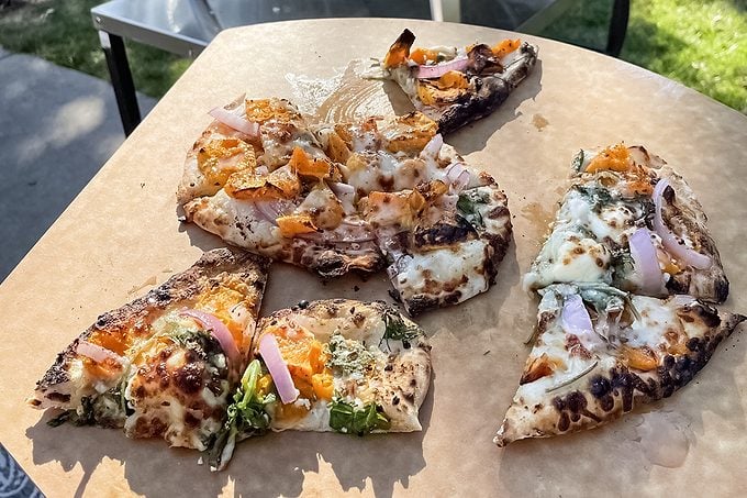 A close-up of a rustic pizza cut into uneven slices, topped with cheese, orange bell peppers, red onions, and leafy greens on a brown serving board outdoors.