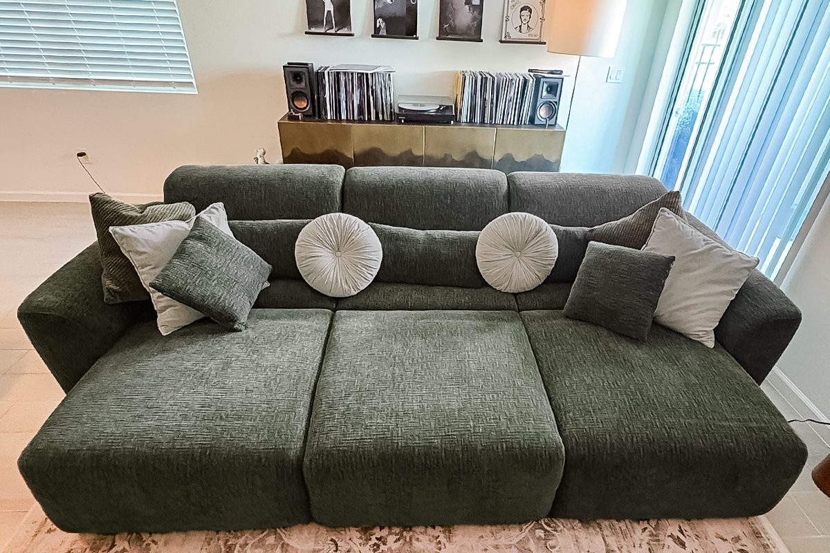 Olive green sofa with six decorative pillows, including round and square shapes, positioned in a modern living room with records and framed photos on a console behind it. Light streams in from a window on the right.