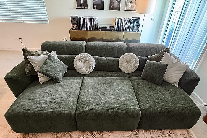 Olive green sofa with six decorative pillows, including round and square shapes, positioned in a modern living room with records and framed photos on a console behind it. Light streams in from a window on the right.