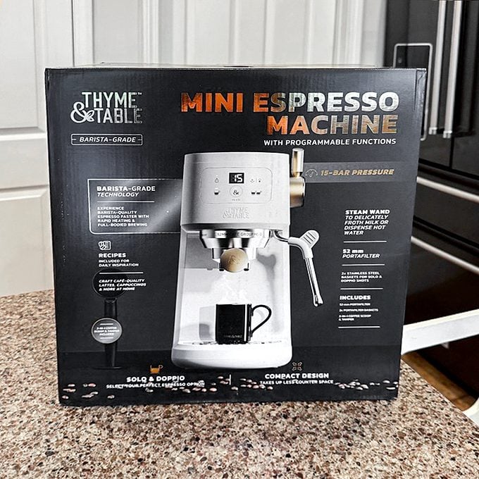 A box containing a Thyme & Table mini espresso machine sits on a kitchen counter. The box is black with orange and white text, featuring an image of the espresso machine and details about its features.