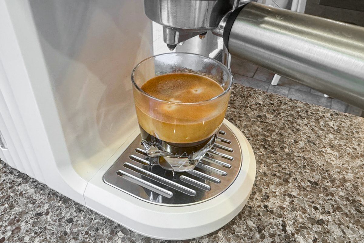 A glass cup filled with freshly brewed espresso sits on the drip tray of a white espresso machine on a speckled countertop.
