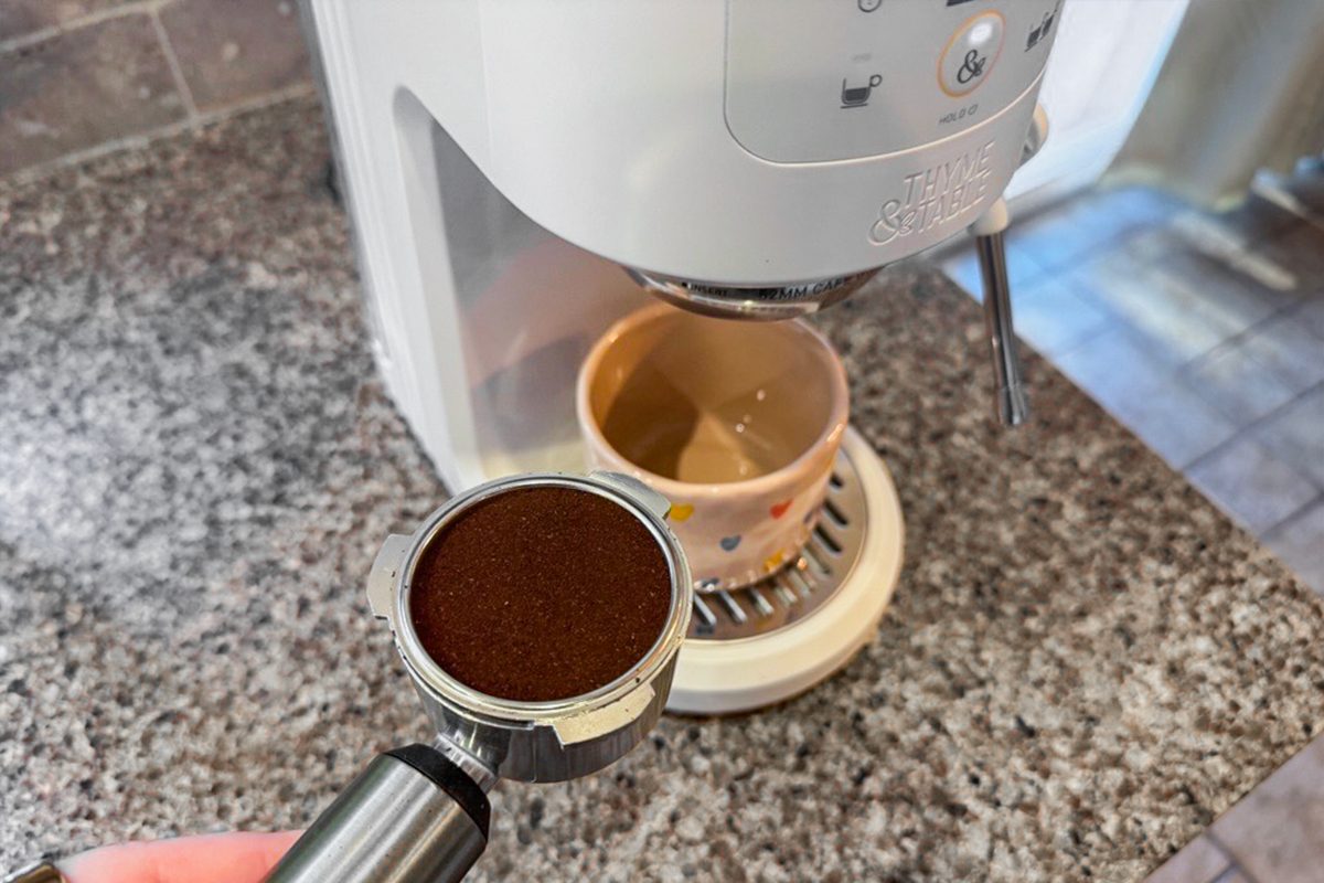 A hand holds a portafilter filled with ground coffee in front of an espresso machine, with a mug placed under the machine’s spout on a granite countertop.