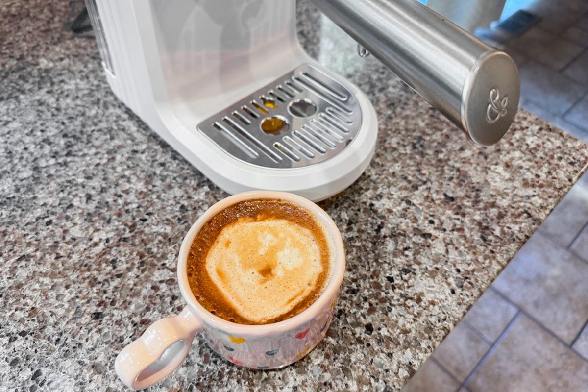 A white coffee machine sits on a granite countertop next to a mug filled with freshly made coffee topped with foam.