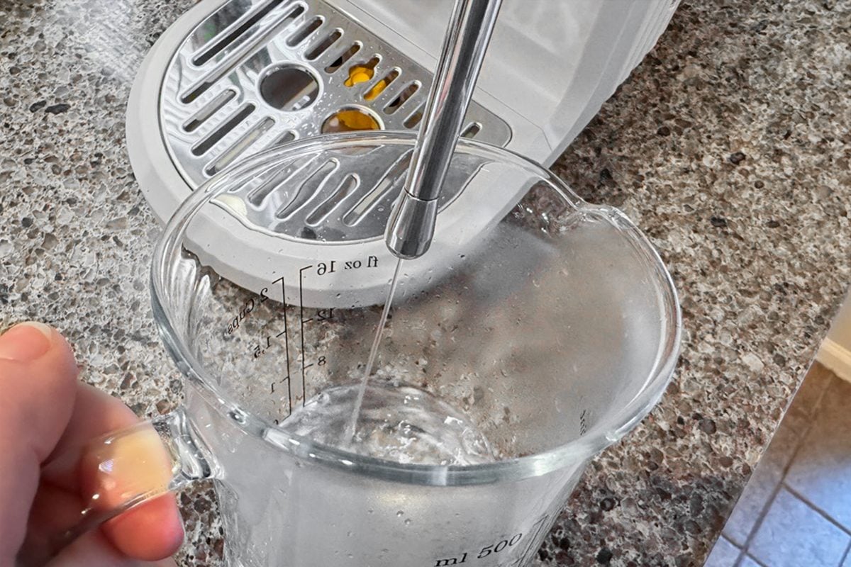A hand holds a glass measuring cup under a steam wand on a coffee machine, with steam entering the cup. The setup is on a granite countertop.