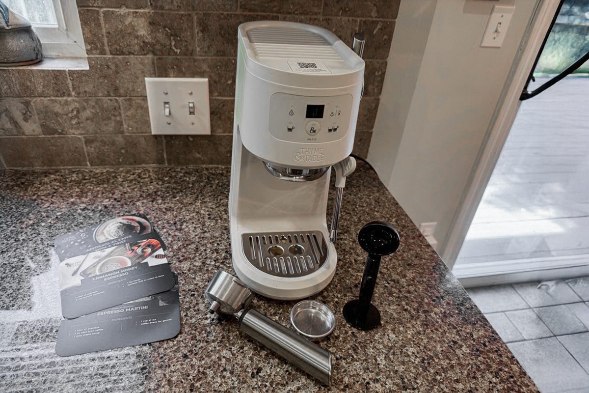 A white espresso machine sits on a kitchen counter with accessories, including a portafilter, filter basket, tamper, and manuals placed nearby. Tile backsplash and electrical outlets are visible in the background.
