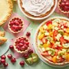 Assorted baked desserts and fresh fruit on a green surface, including a fruit tart topped with kiwi and strawberries, mini cherry tarts, hand pies, a pie with cream topping, and loose cherries, apples, and strawberries.