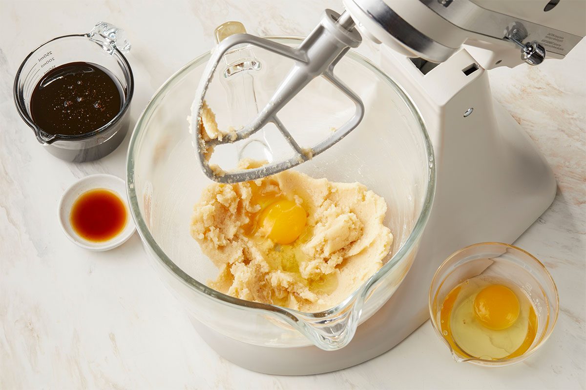 Overhead horizontal image of ingredients being added to the mixing bowl of a stand mixer. Raw eggs, molasses, and vanilla extract are staged around the mixer bowl. | [Original] Creamed butter and sugar in bowl (or KitchenAid). Show adding eggs 1 at a time. Edge in molasses and vanilla.