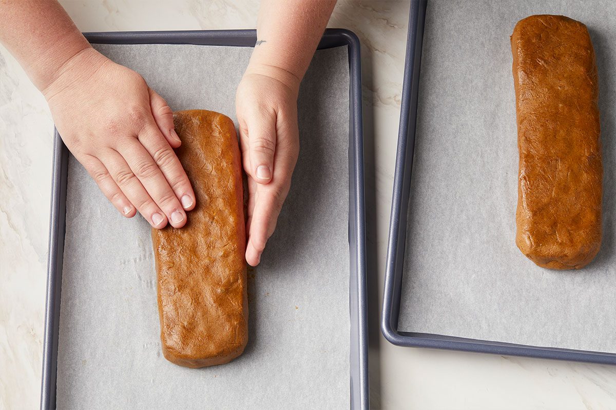 Overhead horizontal image of a pair of hands pressing down on a loaf of cookie dough on a baking sheet lined with parchment paper. A second loaf of dough sits on another baking sheet. | [Original] Divide dough in half. Show one half shaped into a 9x3” rectangle.