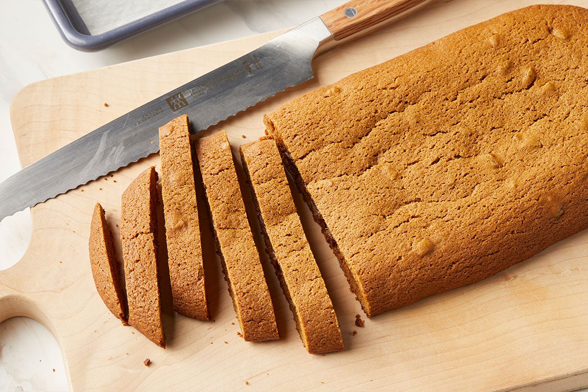 Overhead horizontal image of biscotti being sliced with a ZWILLING J.A. Henckels bread knife on a wooden cutting board. A baking sheet lined with parchment paper sits in the background. | [Original] Cut one log into ½” slices after baking. Show the other sliced and on baking sheet with cut side down.