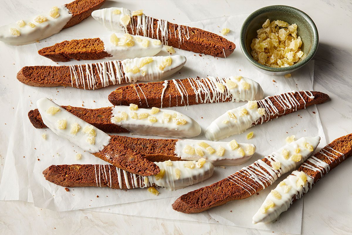 Overhead horizontal image of biscotti. Many biscotti are seen, some dipped in white chocolate with candied ginger. A bowl of candied ginger is also visible.