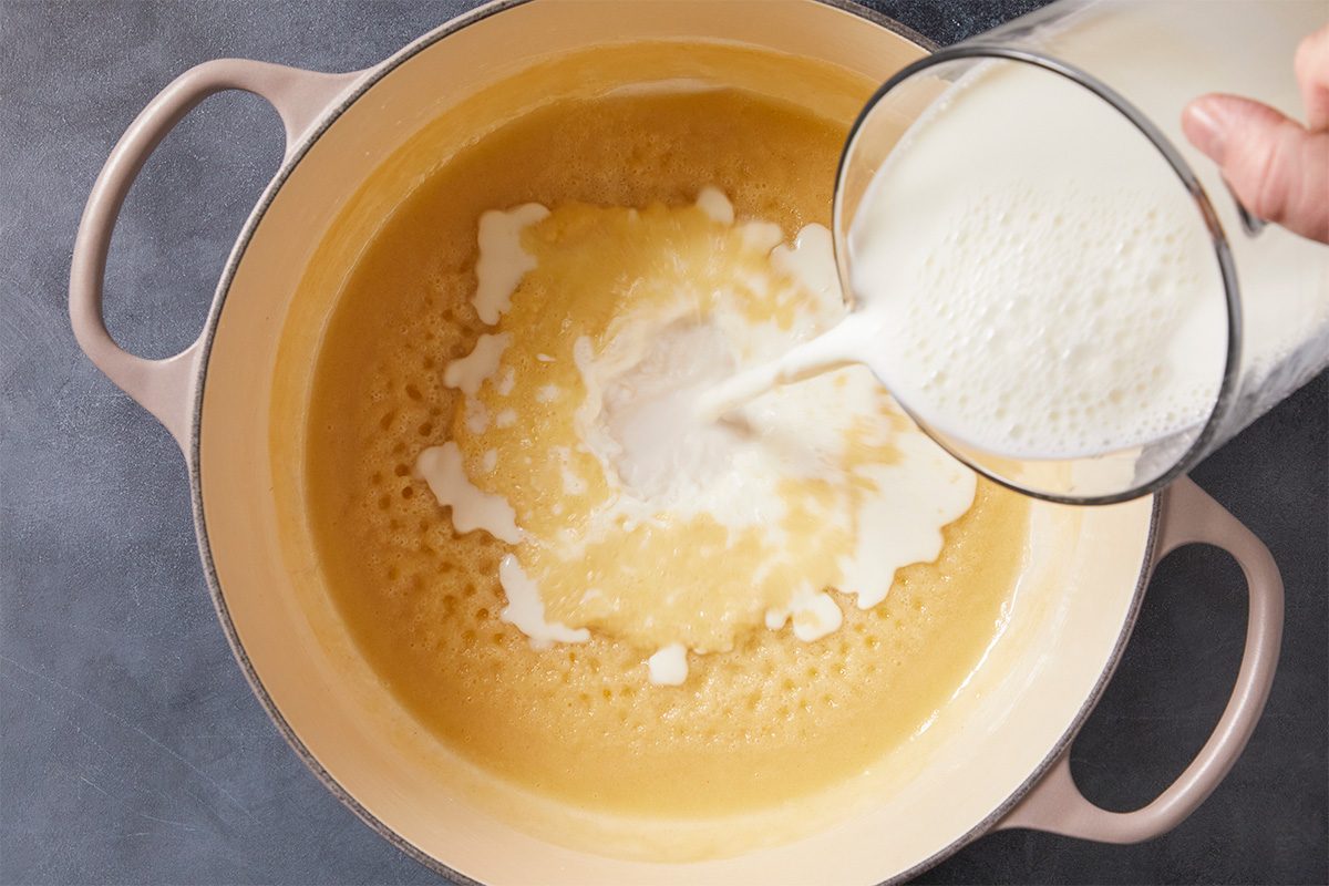 Overhead horizontal image of milk being poured from a glass pitcher into a pot with a light yellow, bubbling liquid. The liquids are combining inside the pot.