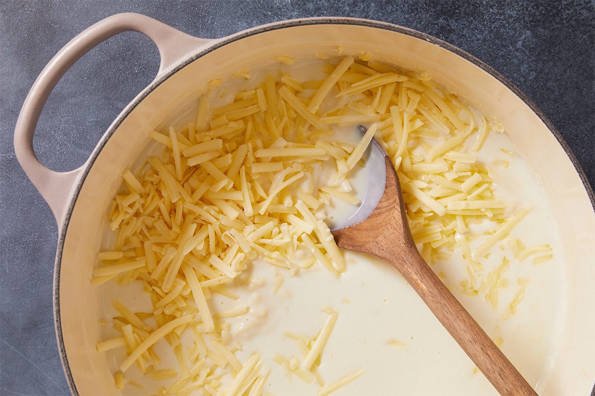 Overhead horizontal image of shredded cheddar cheese being stirred into a creamy white sauce with a wooden spoon inside of a light-colored dutch oven. The pot sits on a dark gray surface.