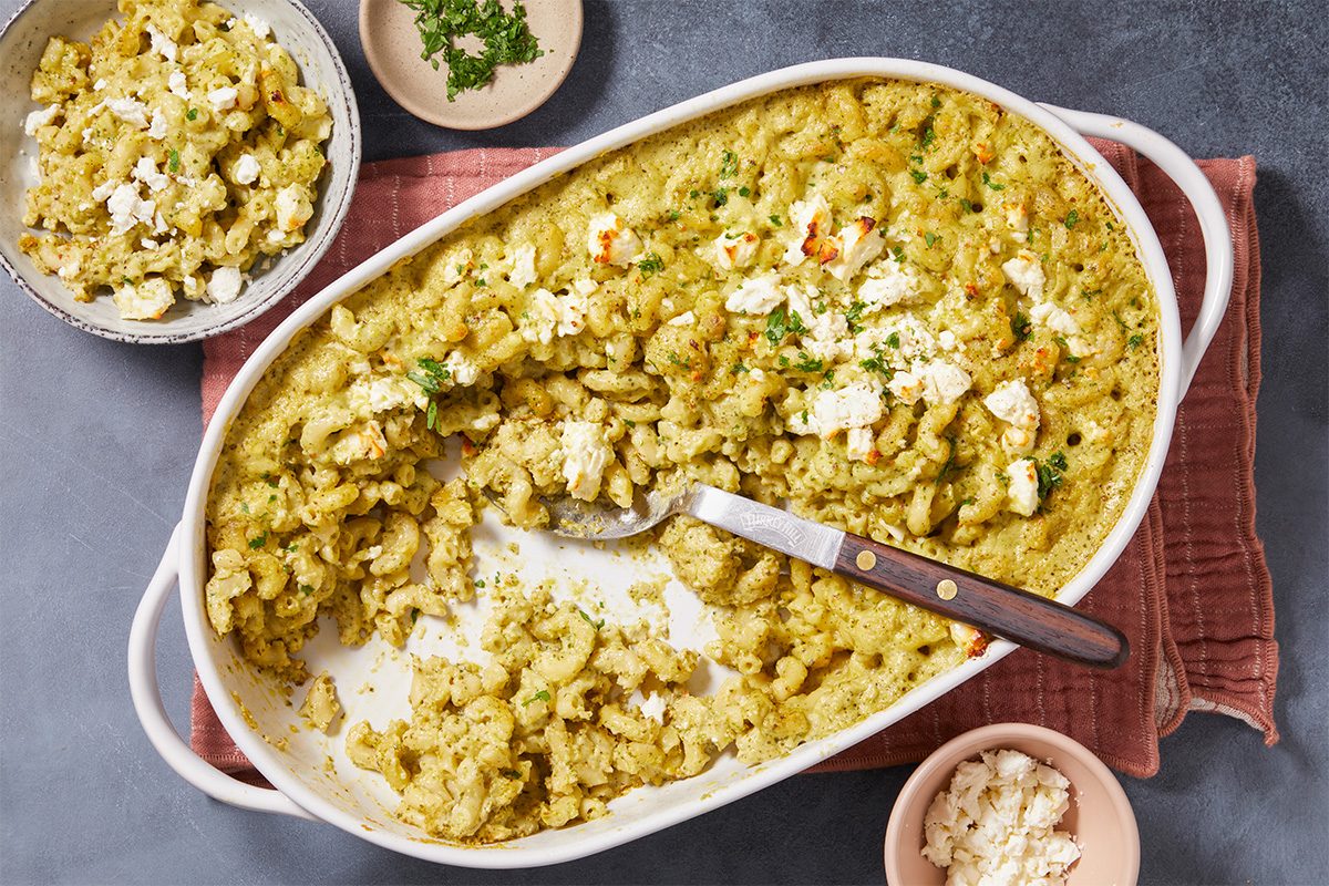 Overhead horizontal image of cheesy pesto pasta bake in a white casserole dish with a spoon scooping a serving. The casserole dish is on a pink linen with a small bowl of feta and some parsley garnish nearby.