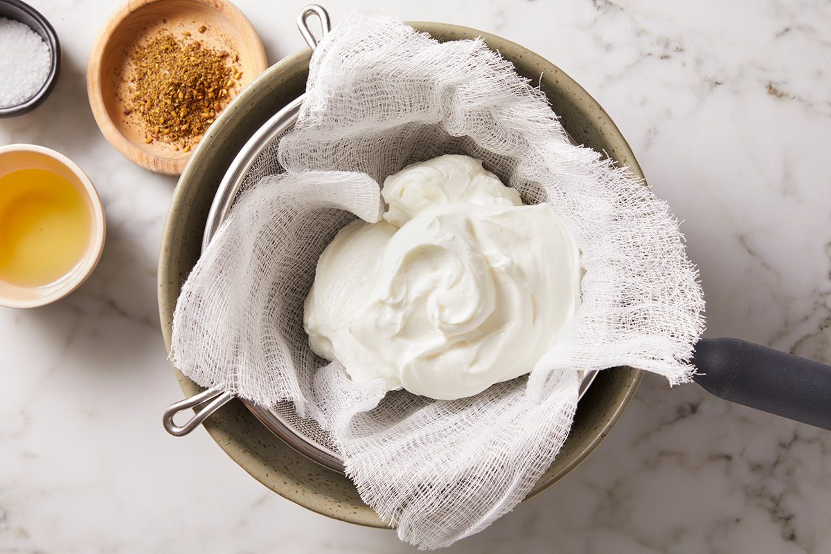 A bowl with a strainer lined with cheesecloth holding thick yogurt, surrounded by a small bowl of olive oil and a dish of brown spices on a marble countertop.