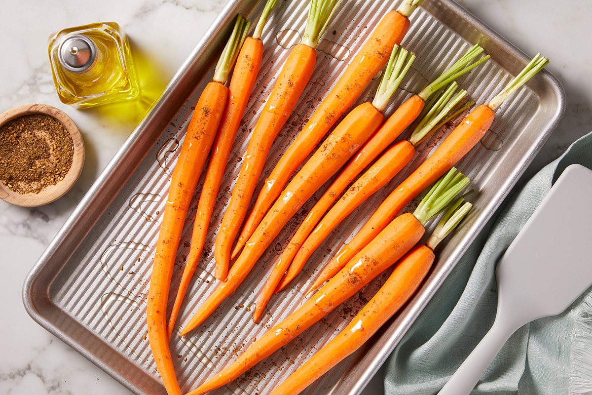 A baking tray holds several whole, peeled carrots with stems, seasoned with herbs and oil. Nearby are a bottle of olive oil, a small bowl of spices, and a white spatula on a marble surface.