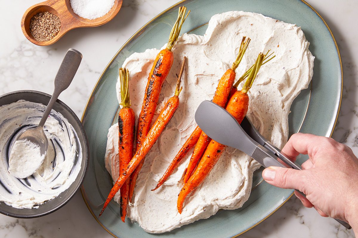 A hand uses tongs to place roasted carrots on a large oval plate spread with a creamy white sauce. A bowl of sauce and a small wooden salt and pepper dish are nearby on a marble countertop.