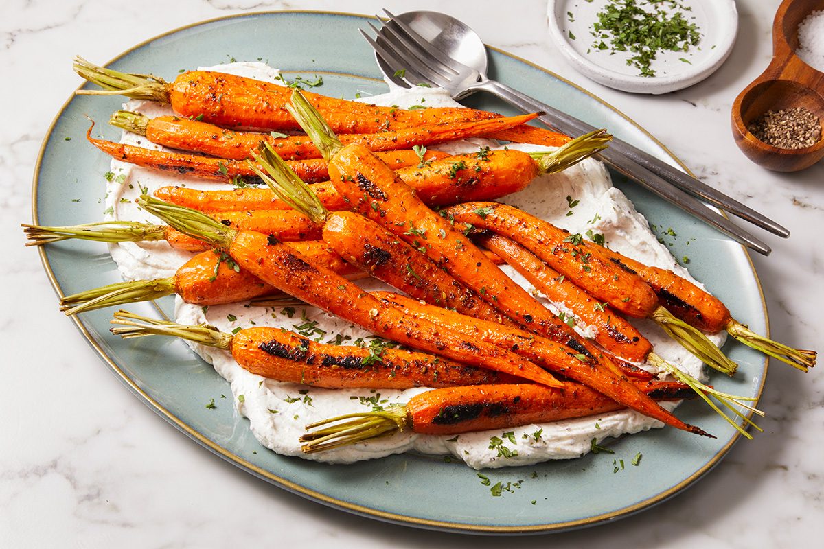 A plate of roasted carrots with green tops, served on a layer of creamy white sauce, garnished with chopped herbs. A fork and spoon rest on the side of the plate.