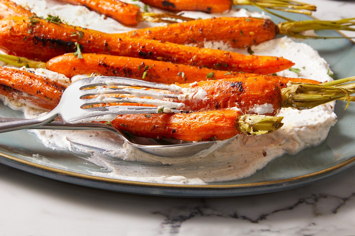 Glazed roasted carrots are served on a layer of creamy white sauce on a plate, with a fork and spoon placed underneath some of the carrots.